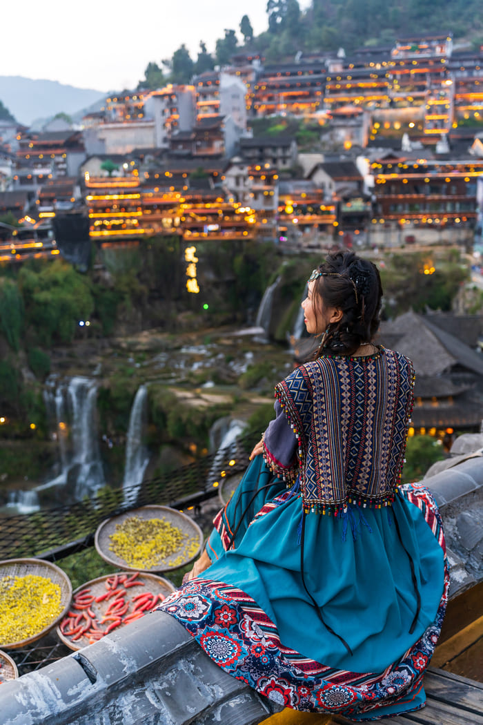 Mujer con vestimenta tradicional contemplando un pueblo histórico iluminado con cascadas, una experiencia clave en nuestros viajes personalizados a China.